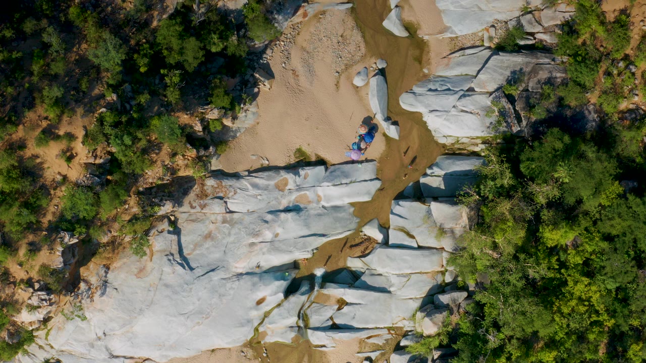 Overhead drone frame from La Palma, Baja California Sur showing pale rock slabs, tan sand, dense green brush, a narrow muddy stream channel, and small colorful figures or gear near the rock edge.