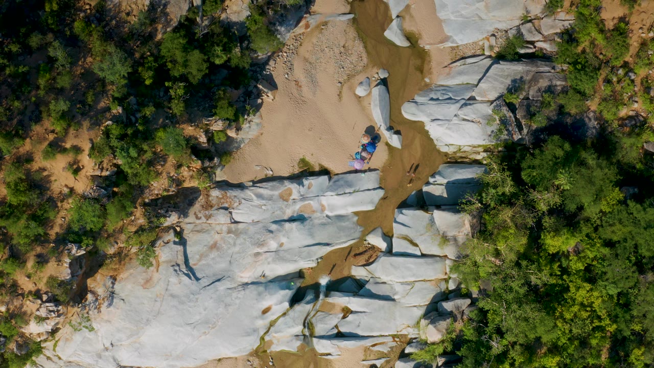 The frame looks straight down over La Palma, Baja California Sur terrain, with pale rock crossing the lower half, sandy wash at the top, green trees on the right, and tiny colorful shapes near the rock that may be people or gear.