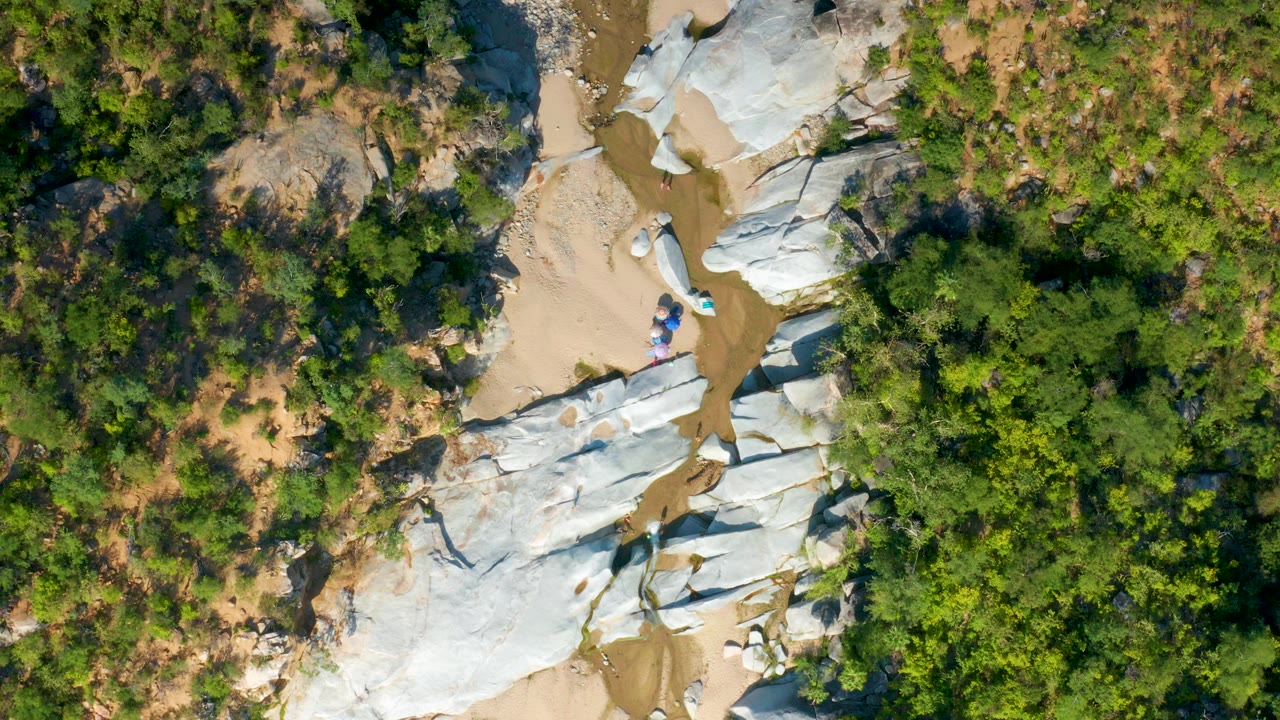 Top-down aerial frame over a dry riverbed in La Palma, Baja California Sur, with pale rock slabs, tan sand pockets, green vegetation, and no visible people.