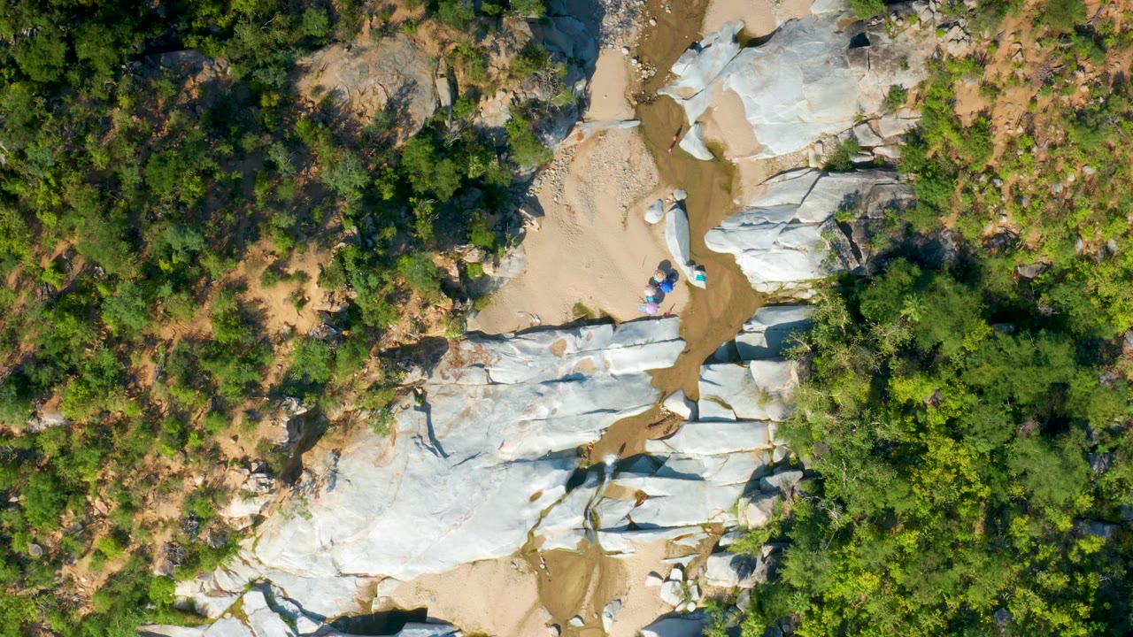 Top-down La Palma frame with the sandy path centered between smooth white rocks, green vegetation along the sides, small blue objects near the wash, and no visible people.