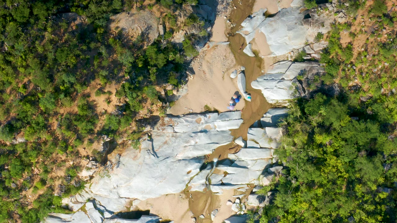 Aerial frame over La Palma terrain showing more green brush above the white rock shelves, tan sand around the channel, a thin muddy stream, and no visible people.