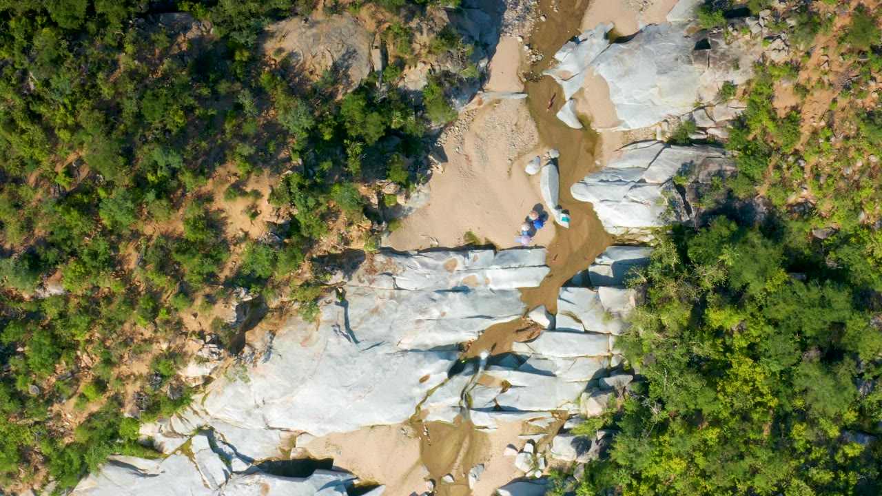 Overhead La Palma view with the sandy wash running past broad white rock surfaces, thick green shrubs at the edge, small blue objects near center, and no visible people.