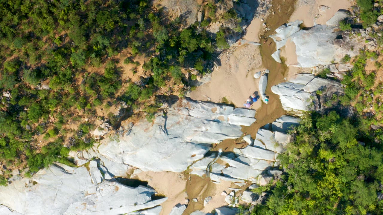 Top-down view of pale rocks, sand, green trees, and a narrow muddy channel.