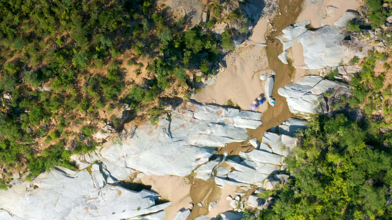 Aerial frame over La Palma, Baja California Sur with white rock outcrops stretching across sandy ground and dense green trees along the upper edge, with no visible people.
