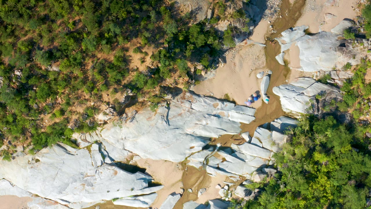 Aerial frame in La Palma, Baja California Sur with white stone formations across the lower area, tan sand through the center, and green tree cover above, with no visible people.