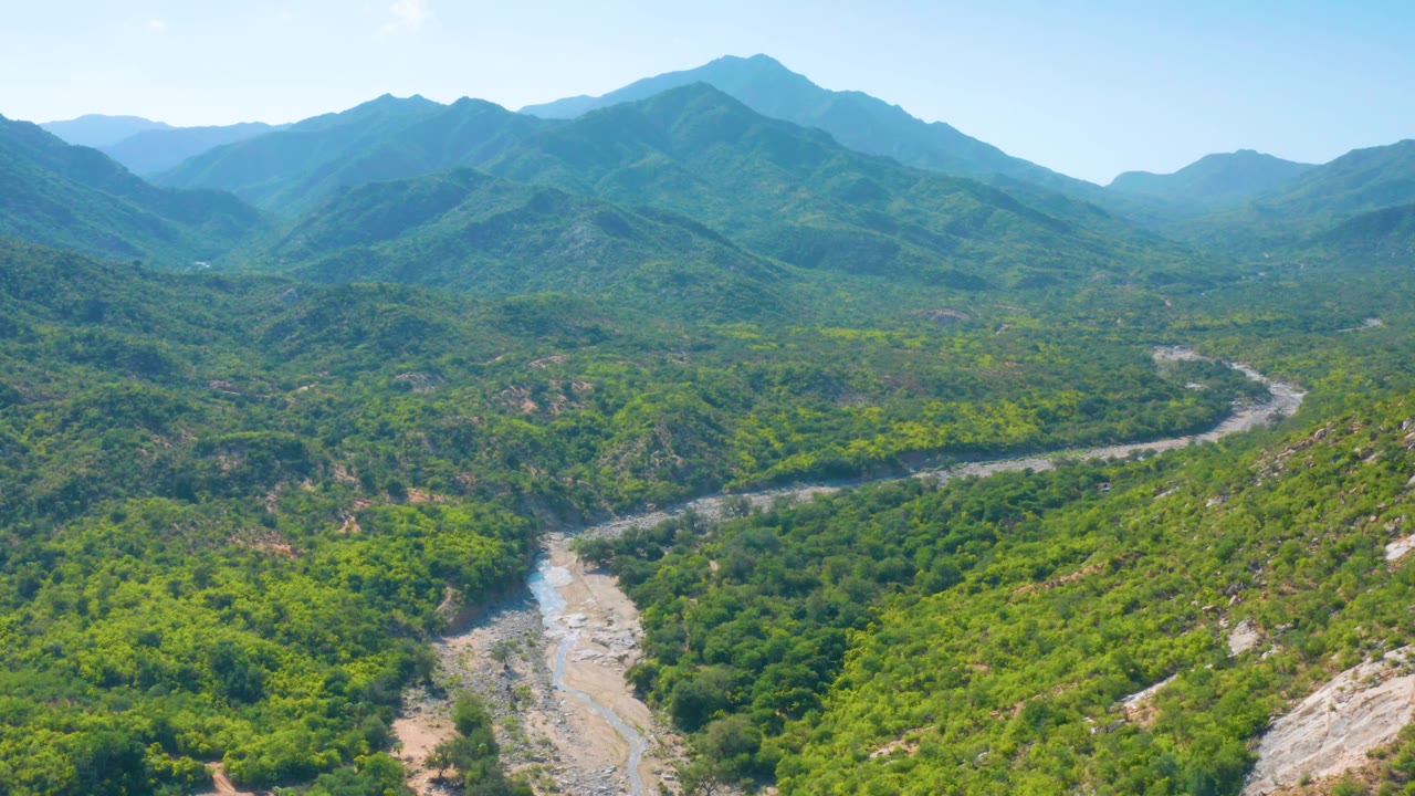 Aerial frame in La Palma, Baja California Sur, showing the nearly dry river wash bending through dense green vegetation below hazy mountain ridges with no visible people.