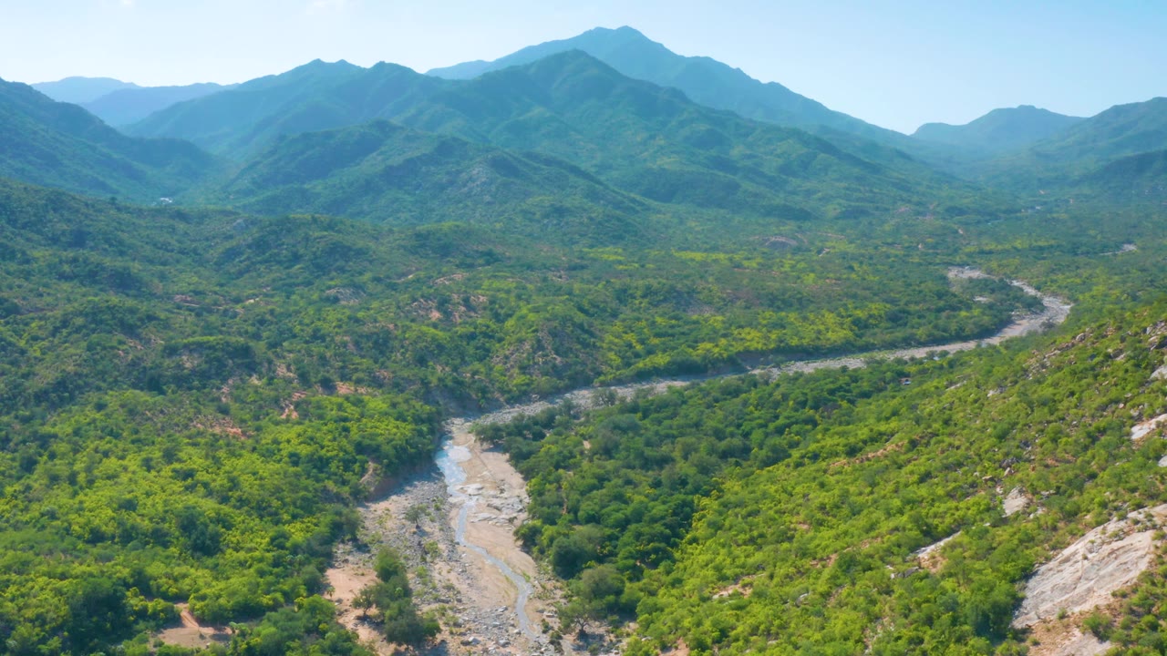 Wide aerial view over La Palma, Baja California Sur, with the dry channel running from the lower frame into green foothills and blue mountain layers beyond, no visible people.