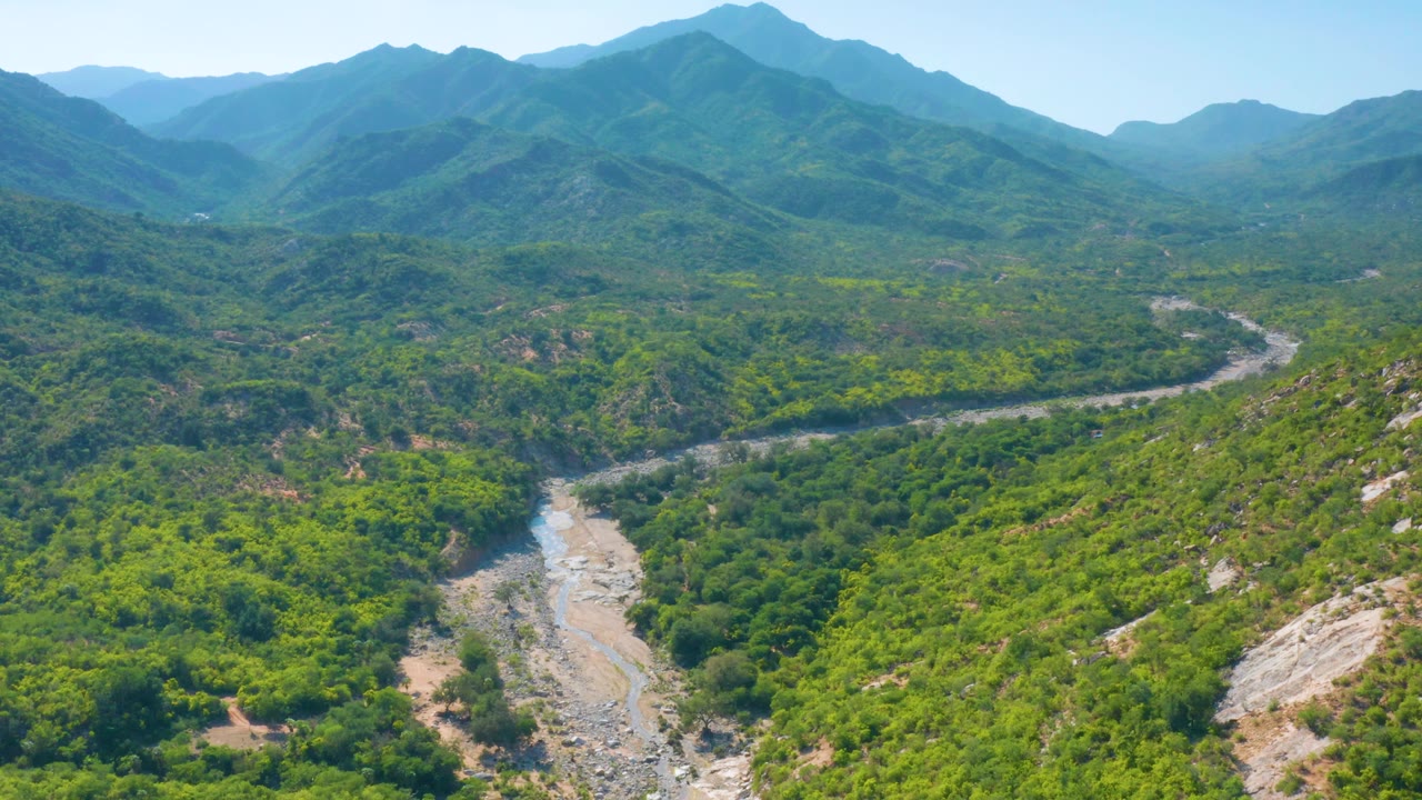 Aerial view in La Palma, Baja California Sur, with a pale stony riverbed in the lower center, green brush across the valley, and mountain ridges filling the background, no visible people.