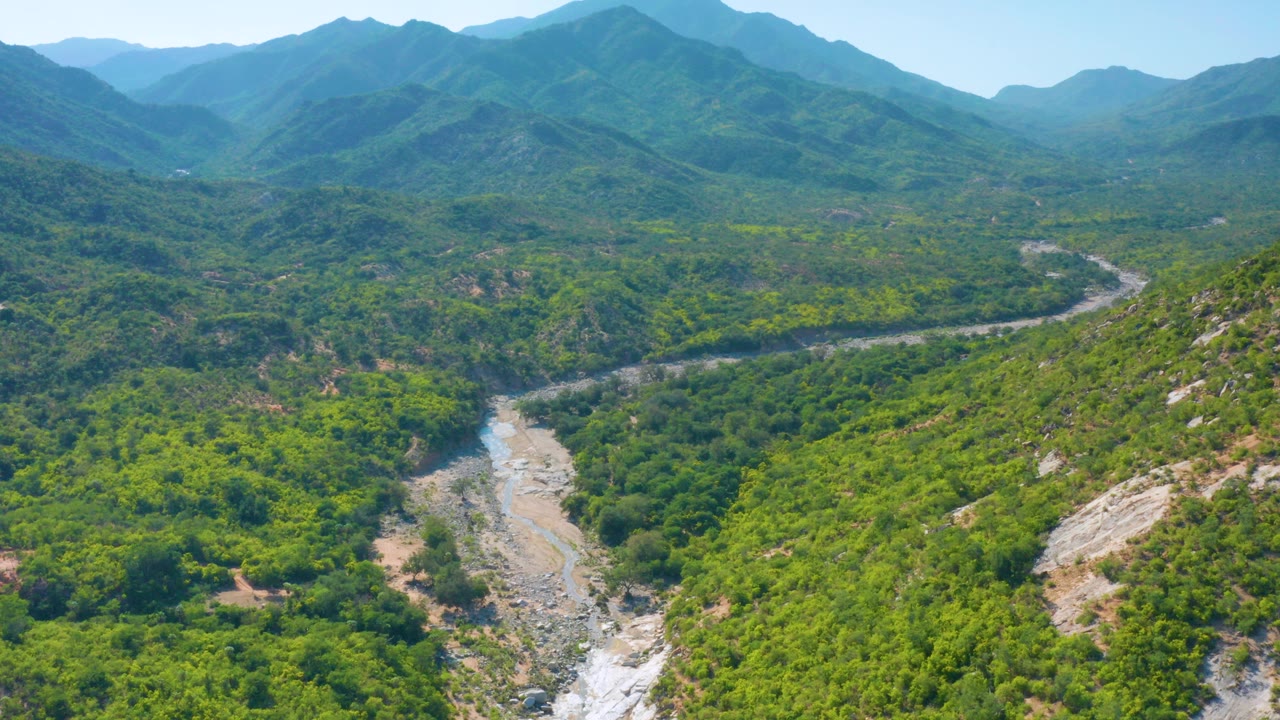 Aerial frame over La Palma, Baja California Sur, showing a rocky riverbed curving through dense green forest below hazy blue mountain ridges, with no visible people.