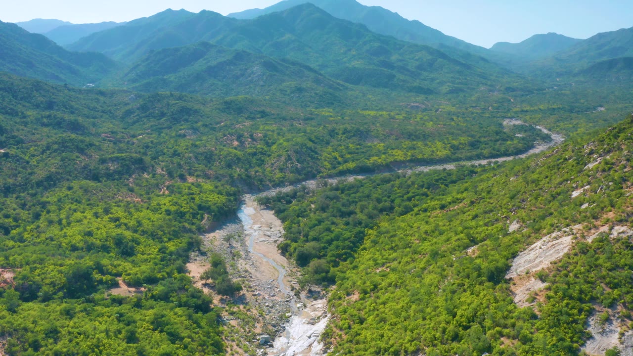 Drone-style aerial view over La Palma, Baja California Sur, with the dry-looking river wash threading between green hills and receding mountain ridges, with no visible people.