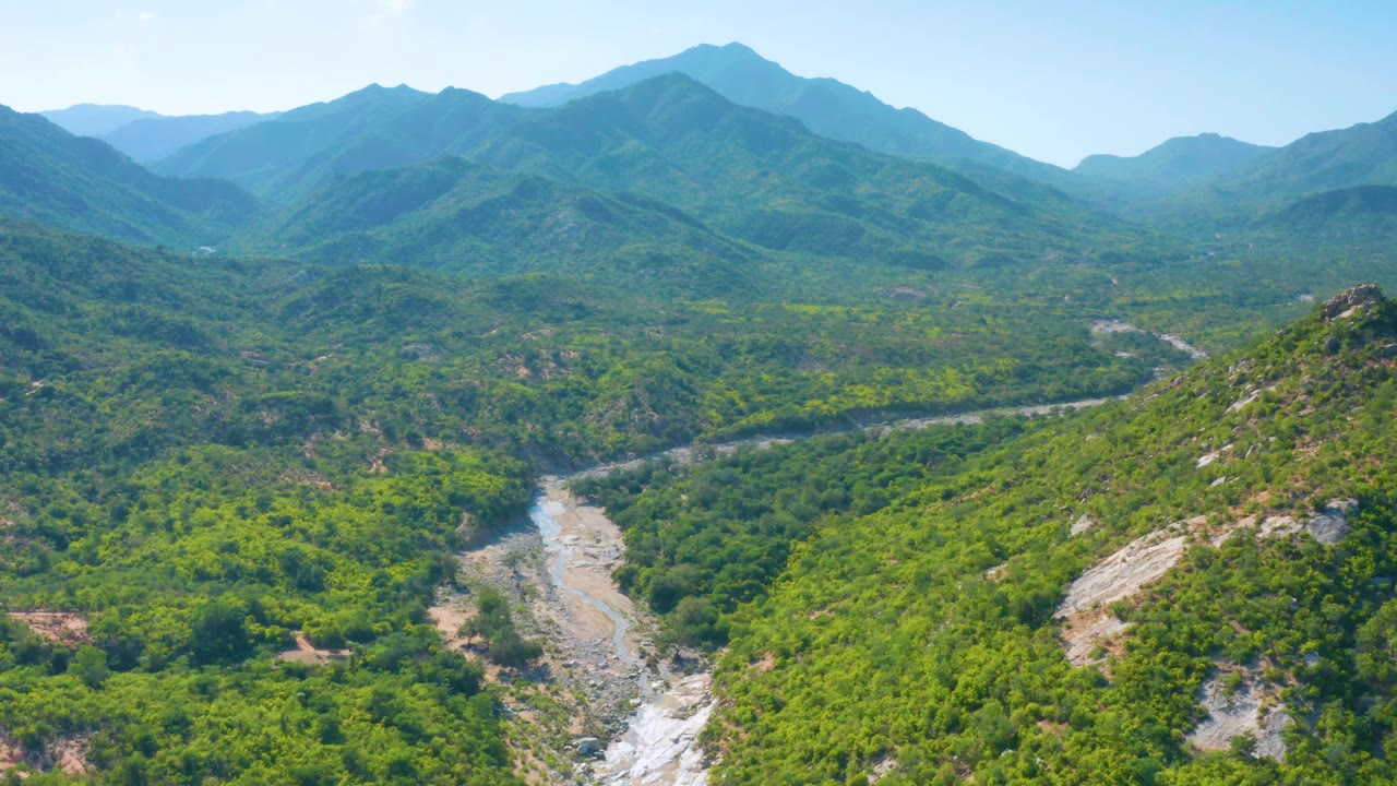 Wide aerial frame in La Palma, Baja California Sur, showing forested hills, a rocky foreground ridge, a light valley corridor, distant mountain layers, and no visible people.