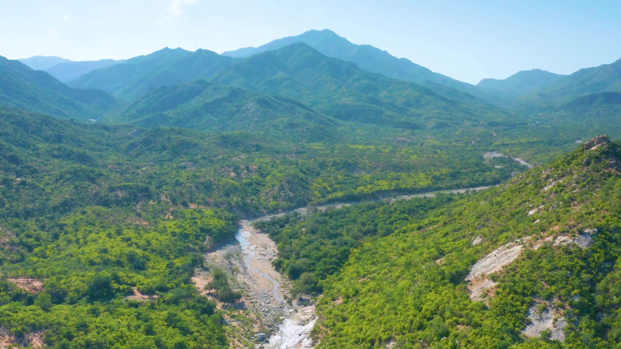 Drone-height view over La Palma, Baja California Sur, with the pale valley route more centered between green hills, layered mountains behind it, and no visible people.