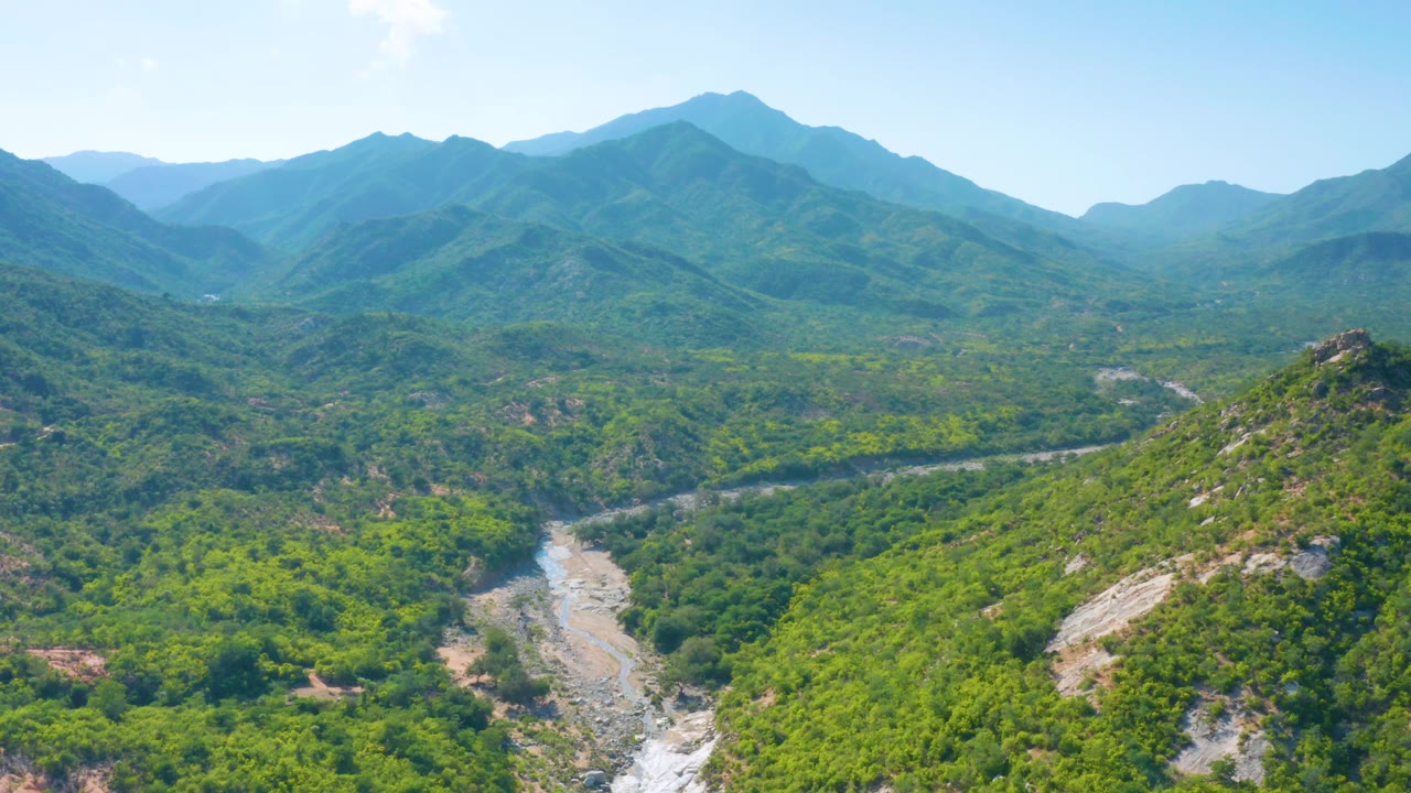 Aerial frame from La Palma, Baja California Sur, looking across dense green hills toward hazy blue mountain ridges, with a pale valley line and no visible people.