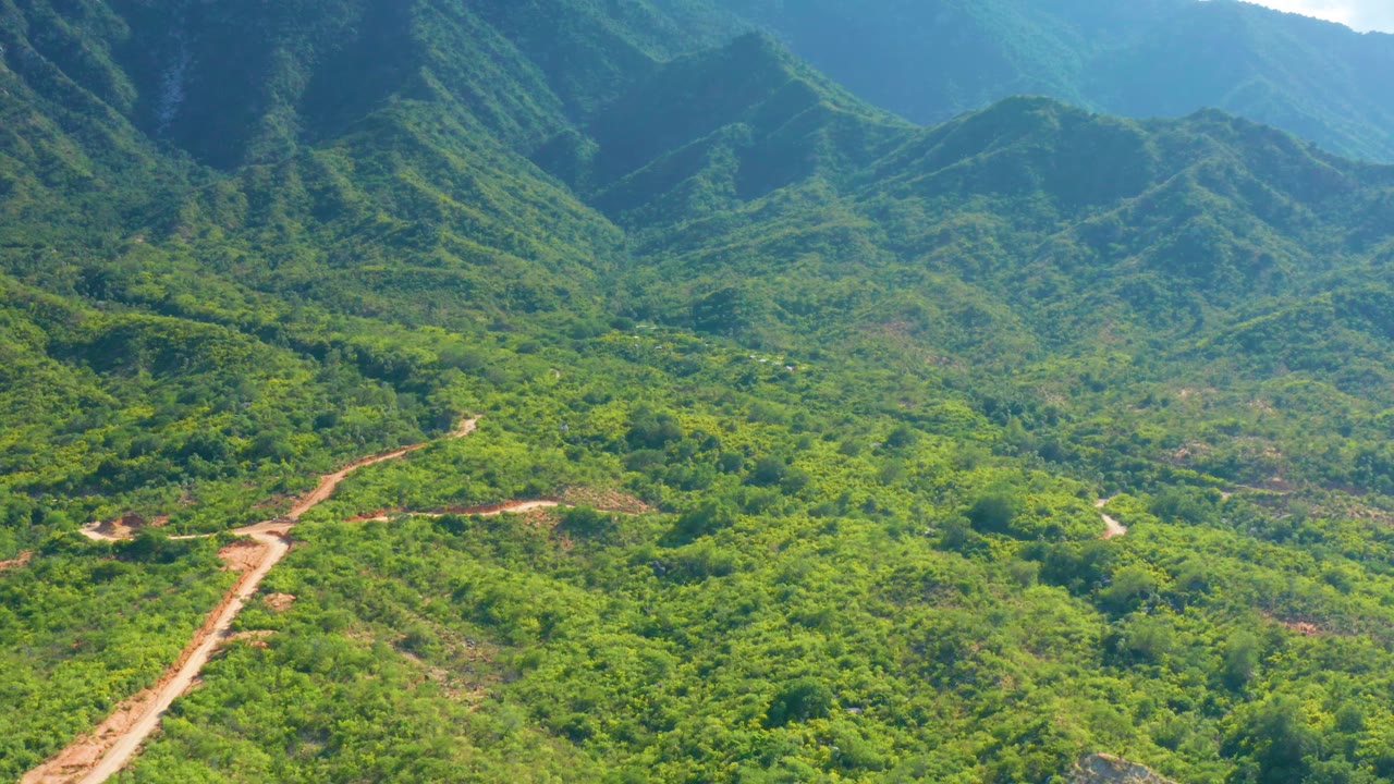 Aerial view over La Palma, Baja California Sur, with a winding mud road cutting across the lower hills and dense green vegetation under bright light, with no visible people.