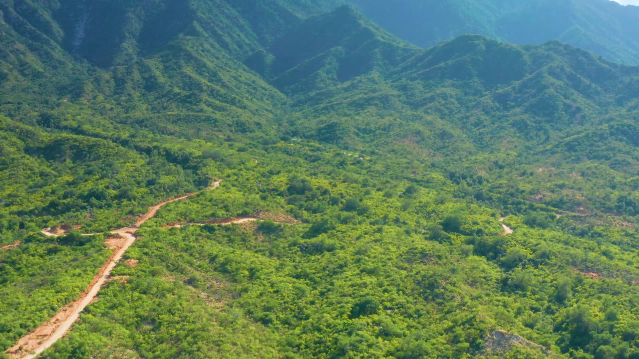 La Palma aerial frame showing the mud road entering from the lower left, scattered bushes across the valley, and steep shaded mountain slopes in the background, with no visible people.