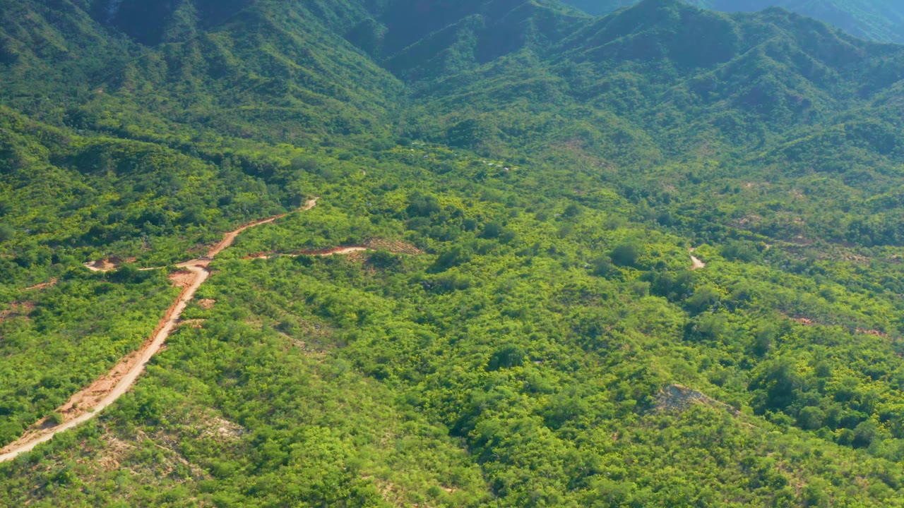 An aerial frame near La Palma, Baja California Sur shows a winding dirt road crossing a green hillside toward layered mountain ridges, with no visible people.