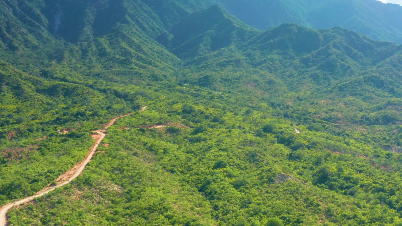 La Palma, Baja California Sur aerial view with the dirt road entering from the lower left edge, green hills spreading across the valley, blue mountain ridges behind, and no visible people.