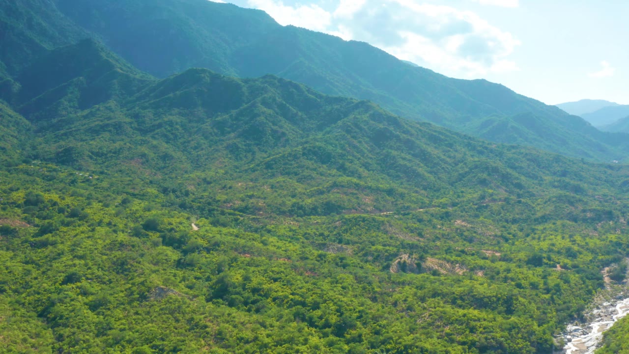 Early aerial frame over La Palma, Baja California Sur, showing forested slopes, layered ridgelines, the rocky river channel near the lower right edge, bright sky, and no visible people.