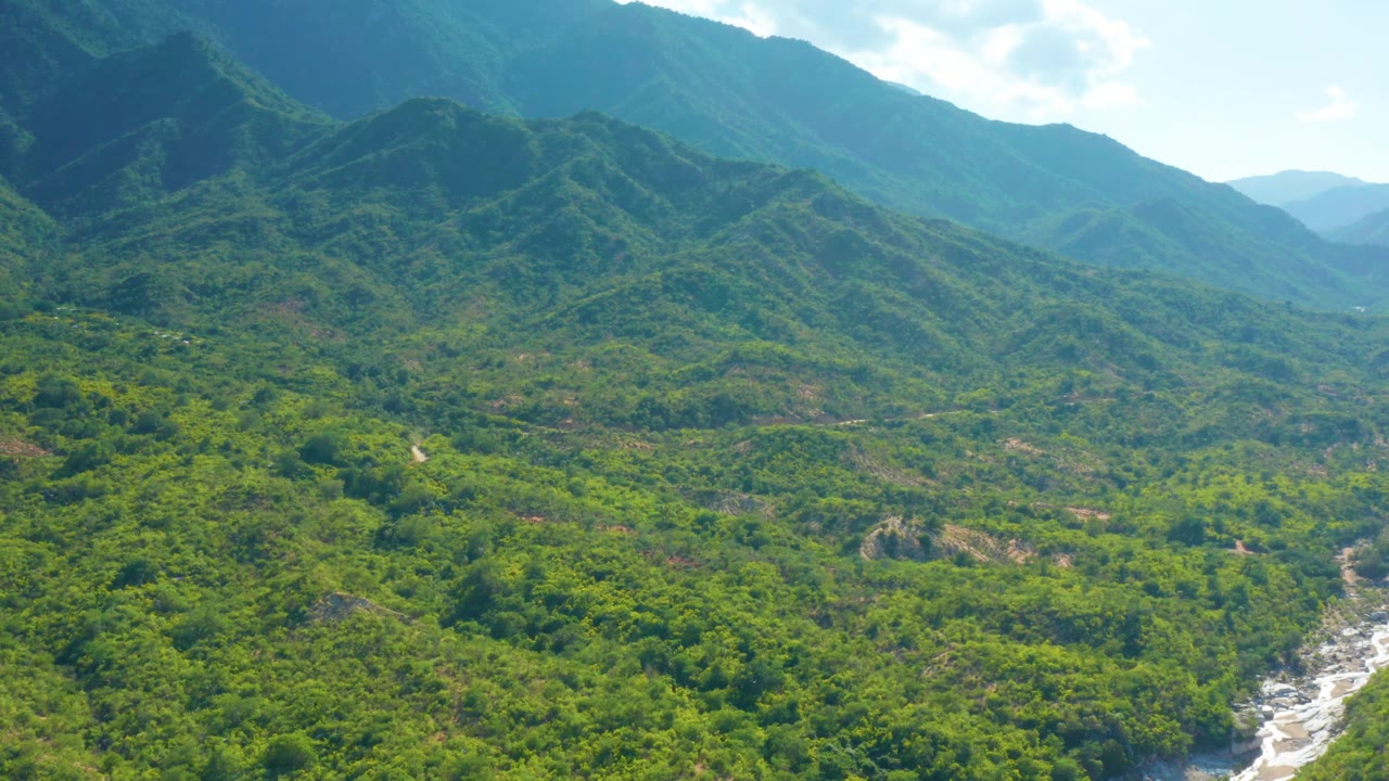 Mid-clip aerial frame over La Palma, Baja California Sur, showing green slopes descending toward a pale rocky river course, darker mountain shade at upper left, distant ridges, and no visible people.