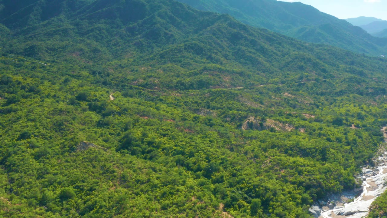 Aerial frame over La Palma, Baja California Sur, showing dense green hills, distant mountain ridges, and a pale rocky creek bed near the lower right with no visible people.