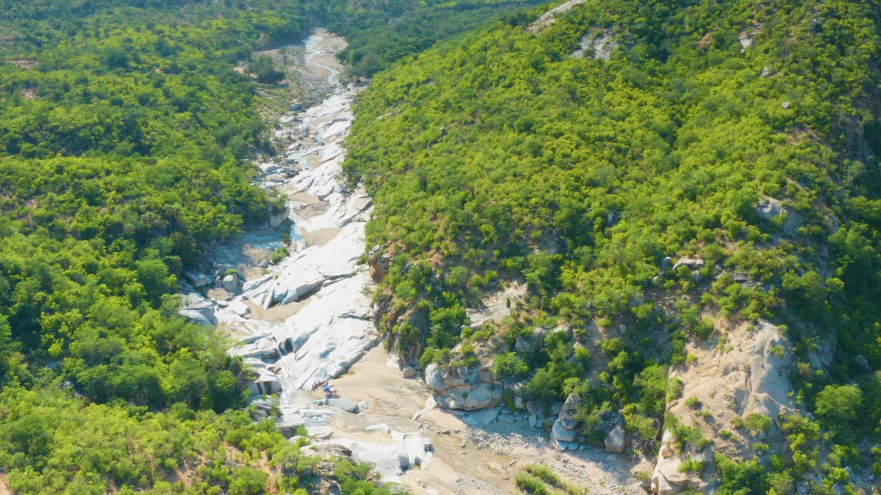 A wide aerial frame in La Palma, Mexico shows the arroyo continuing through a valley of green brush and exposed pale rock, with no visible people.