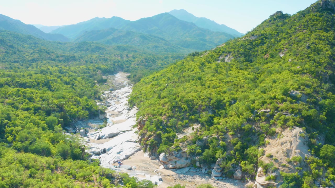 Aerial valley view in La Palma, Baja California Sur, with green slopes framing a light rocky creek bed below blue mountain ridges, no visible people.