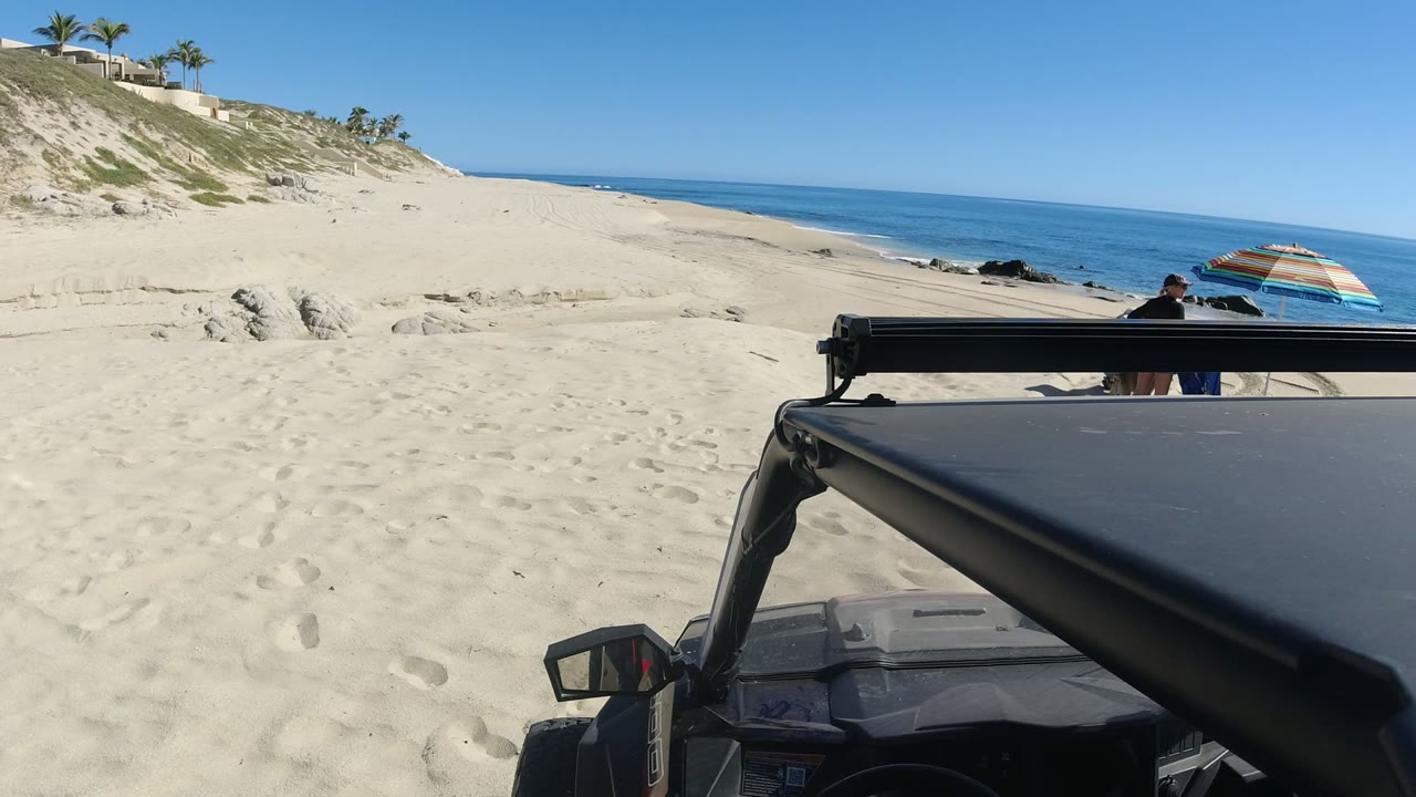 At the start of the Los Cabos clip, a dark off-road vehicle edge fills the lower frame while pale sand, blue ocean, dunes, palms, a beach umbrella, and partially visible people sit beyond it.
