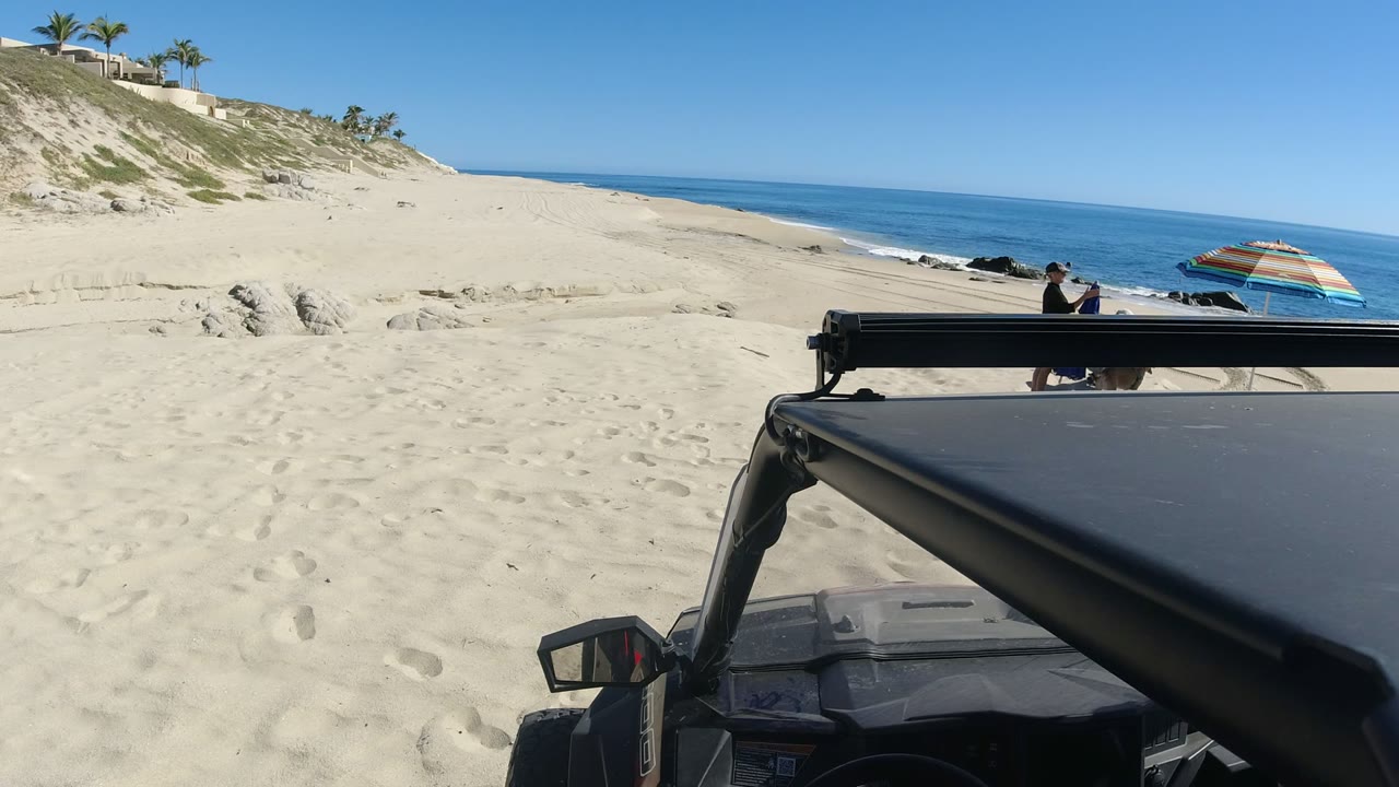 At 0:01 in Los Cabos, Mexico, the vehicle roof and frame remain close to camera as the beach slopes toward blue water and the striped umbrella sits near the right edge.