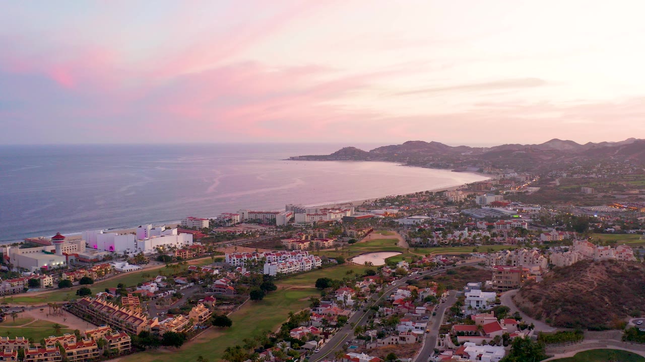 Early frame of San Jose del Cabo from above, with ocean on the left, a curved bay, coastal buildings, hillside homes, distant hills, pink clouds, and no visible people.