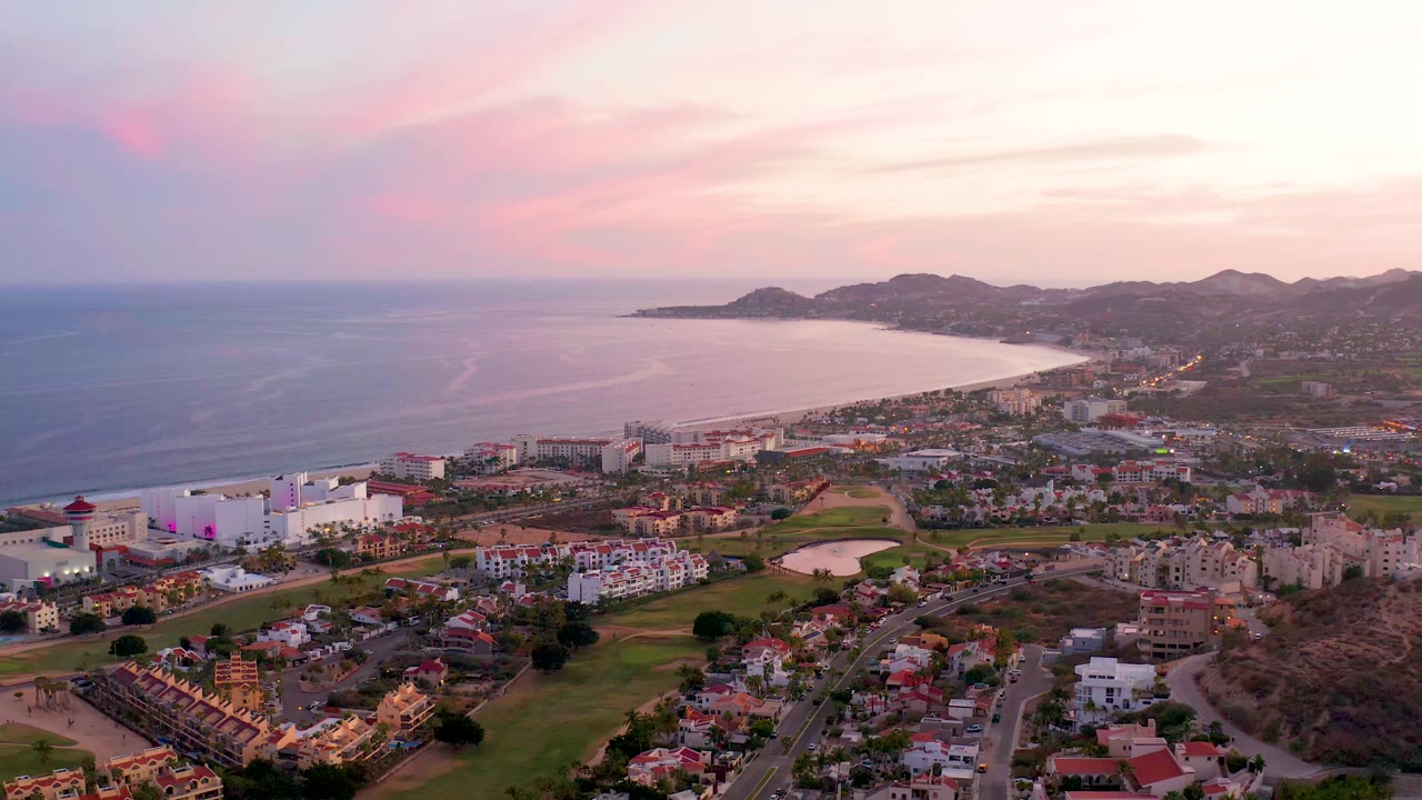 Wide aerial of ocean, beachfront town, grassy area, and hills at dusk.