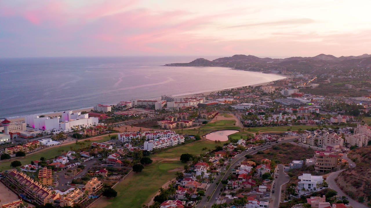 High aerial view over San Jose Del Cabo with the ocean on the left, a curving beach beyond the town, green fairways, roads, homes, distant mountains, pink sunset sky, and no visible people.