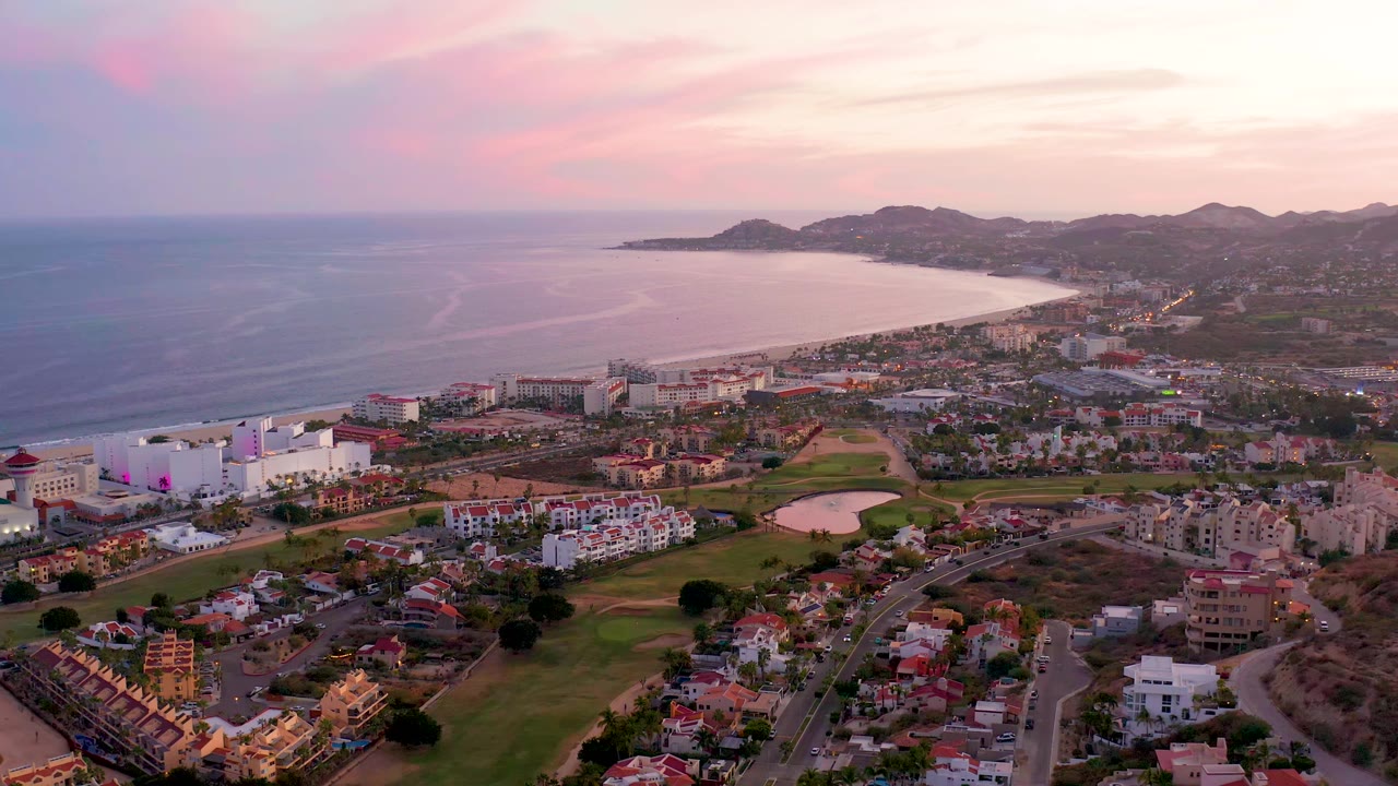Aerial frame of San Jose Del Cabo showing resort-scale buildings near the shore, residential streets and green fairways below, a pale beach curve, calm ocean, mountain horizon, and no visible people.