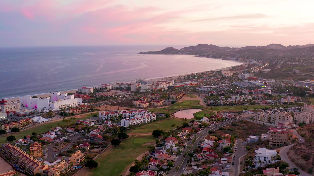 San Jose Del Cabo aerial view with faint surf lines on the left, a long beach running toward the mountains, town blocks and roads below, green fairways in the foreground, pastel sunset sky, and no visible people.