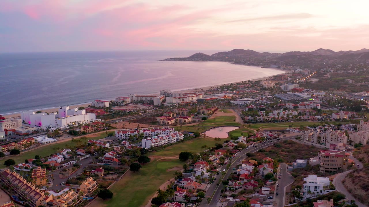 Aerial view of ocean, beach, coastal development, roads, fairways, homes, and sunset mountains.