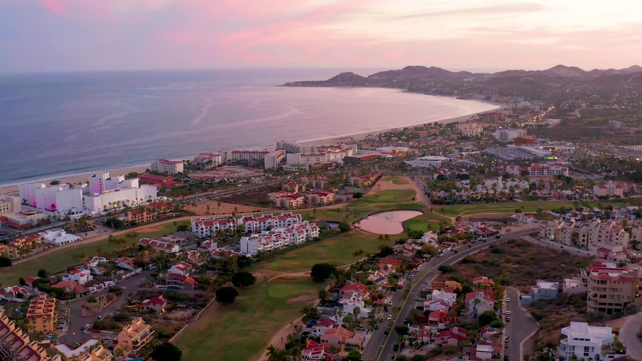 Aerial view of San Jose del Cabo, Mexico, one second into the clip, with the curved beach and ocean on the left, dense low-rise buildings below, distant hills, soft pink sky, and no visible people.