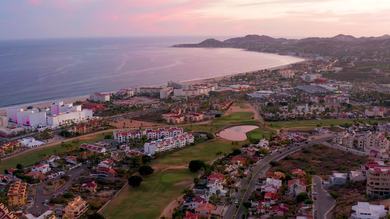 Dusk aerial over coastal buildings, green grounds, curving roads, beach, ocean, and hills.