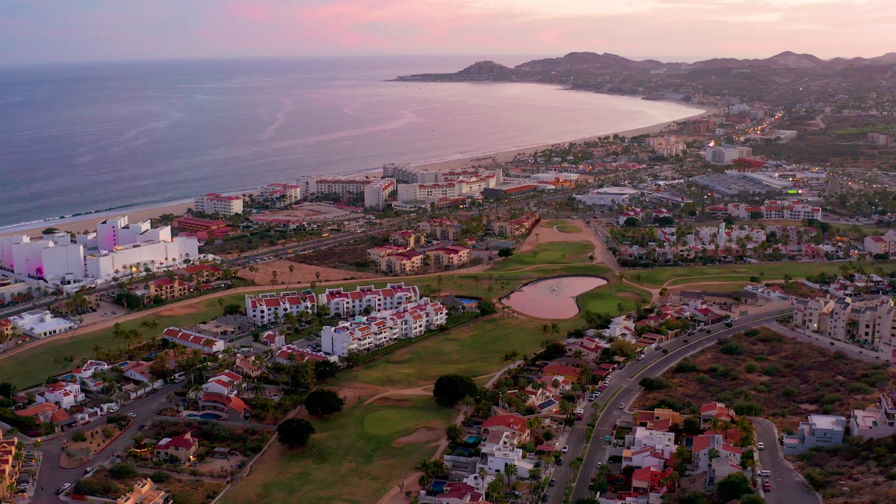Aerial frame over San Jose del Cabo, Mexico, showing the ocean and curving beach behind low-rise coastal buildings, roads, golf greens, and red-roof homes, with no visible people.