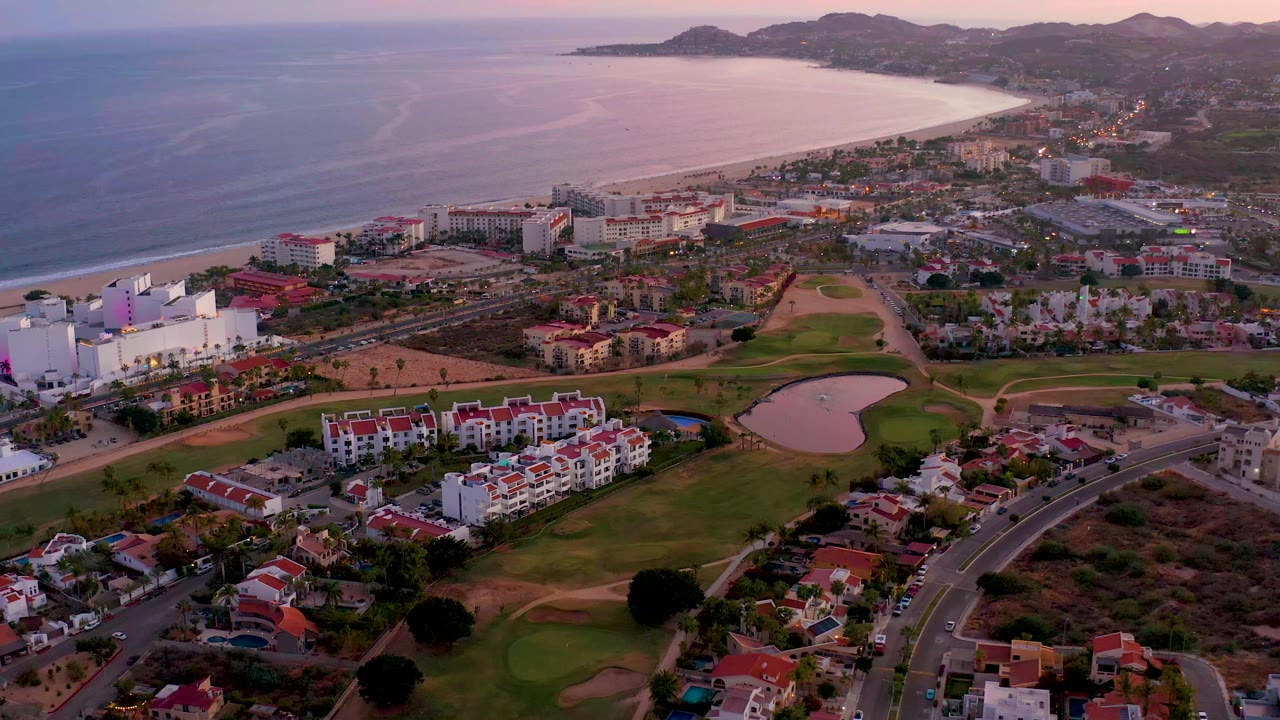 High aerial view in San Jose del Cabo, Mexico with the shoreline stretching across the background, fairways and a pond below, resort-style buildings near the beach, and no visible people.