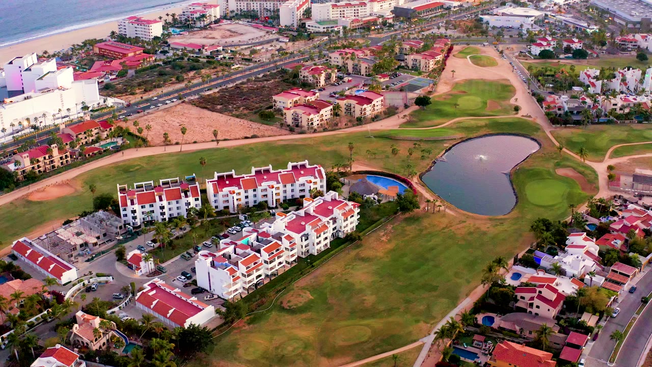 San Jose del Cabo aerial frame with the golf pond centered right, white residential blocks with red roofs below, coastal buildings and beach visible near the top, and no visible people.