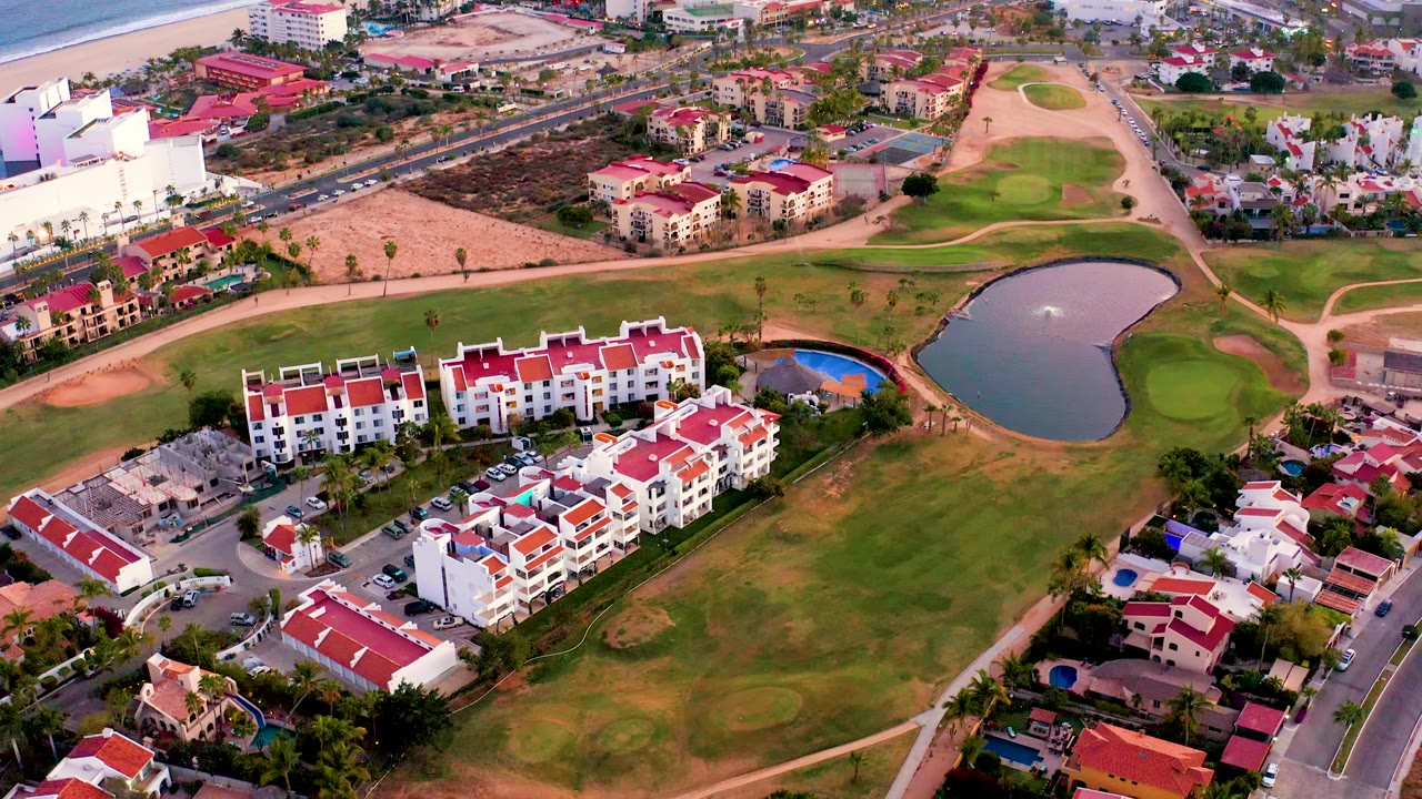 Aerial view of red-roofed residences beside green golf course and pond with roads and beach beyond.
