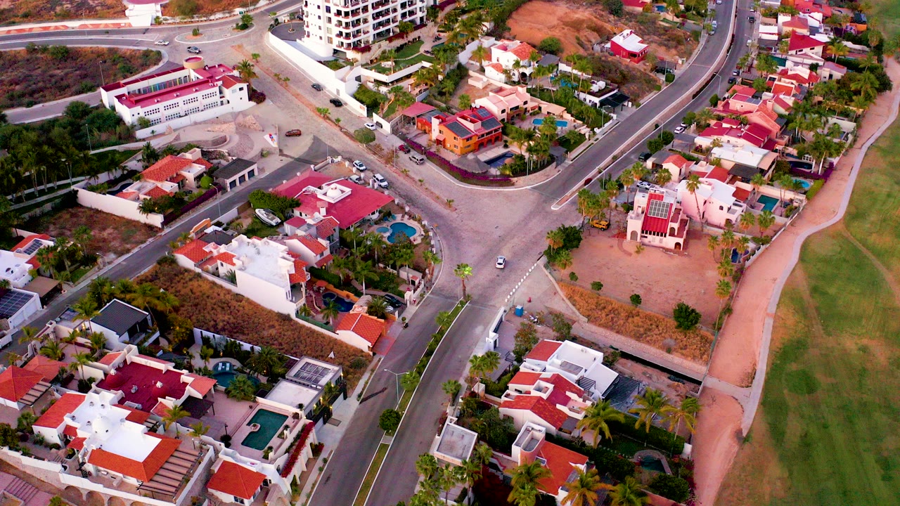 Aerial view over San Jose del Cabo homes with red roofs, a curved intersection, a green golf course edge on the right, and no visible people.