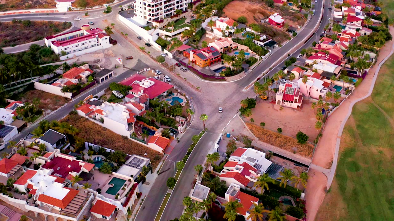 San Jose del Cabo aerial frame showing the intersection centered between homes, a fairway along the right side, parked and moving cars, and no visible people.