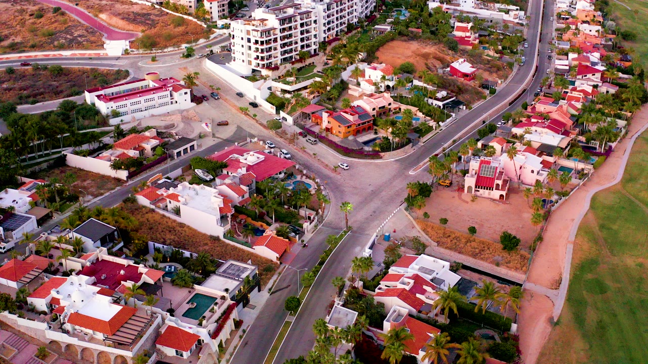 Aerial frame from San Jose del Cabo showing red-roofed homes, winding roads, palm trees, a green fairway-like strip, and a brown hillside with a pink path, with no visible people.