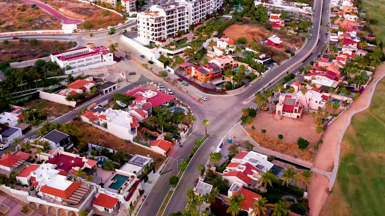 San Jose del Cabo aerial view with foreground villas, a curving road junction, hillside homes, and a white multi-story building near the top of frame, with no visible people.