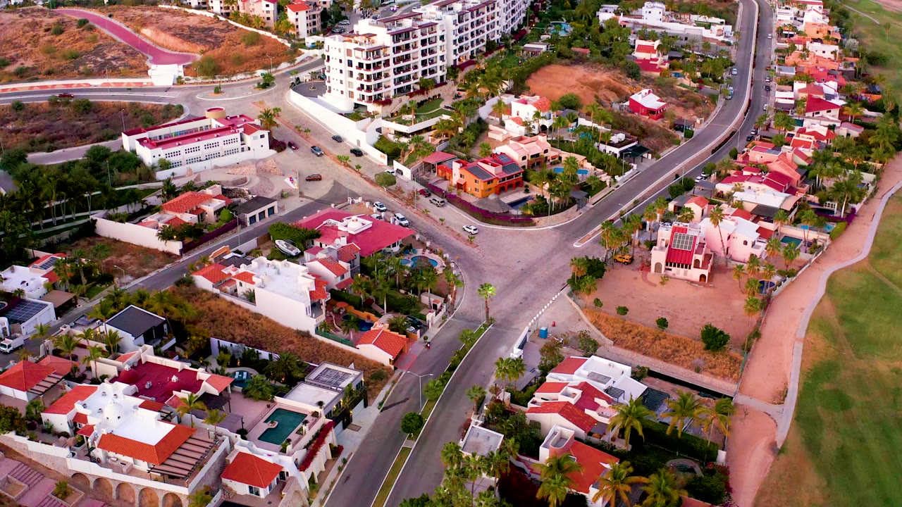 Aerial frame over San Jose del Cabo residential blocks with a sweeping road, clustered red roofs, palms, a green strip at right, and dry hillside above, with no visible people.