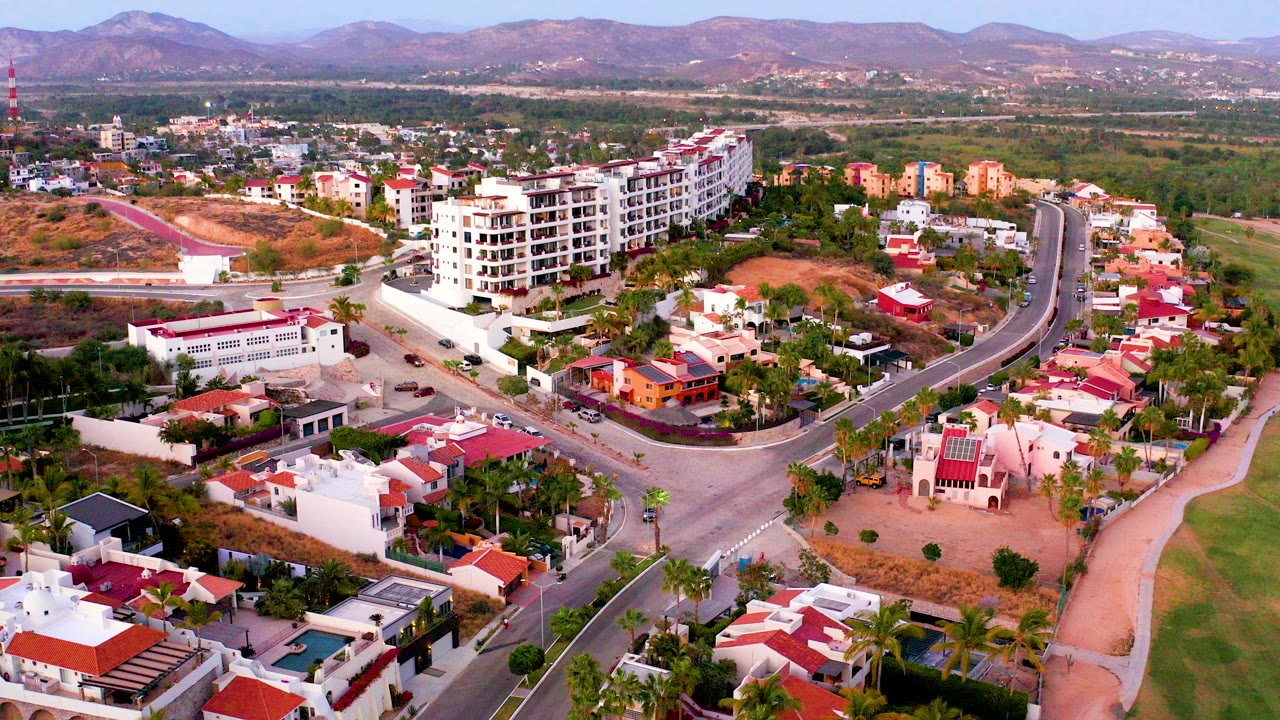 Aerial view over a San Jose del Cabo residential neighborhood with red-roofed homes, a curved road, white apartment buildings, palms, distant mountains, and no visible people.