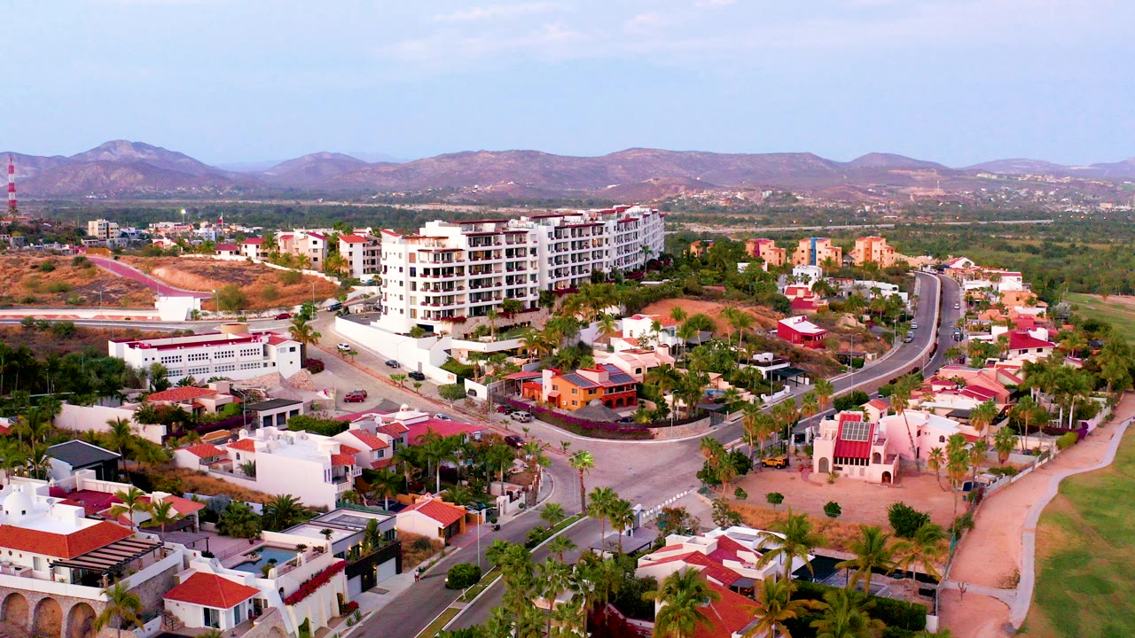 Opening aerial frame over a San Jose Del Cabo residential neighborhood with white buildings, red roofs, palms, curved roads, dry hills, distant mountains, and no visible people.