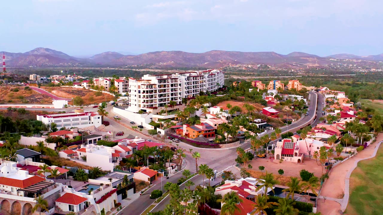 Residential aerial with a curved road, palms, white buildings, and mountain horizon.