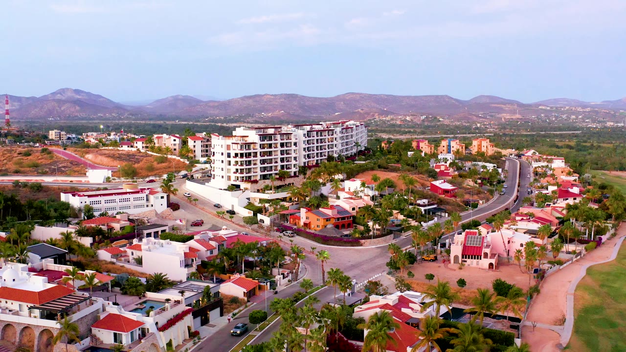Wide San Jose Del Cabo aerial frame with clustered white homes, red roofs, palms, a road network, green lowlands, hazy mountains, and no visible people.