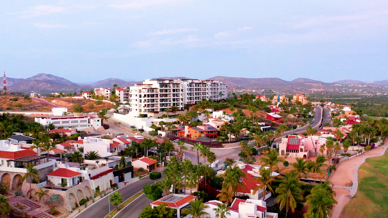 Opening frame of San Jose del Cabo BCS Mexico from above, with a palm-lined road, white homes with red roofs, dry hills, distant mountains, pale sky, and no visible people.